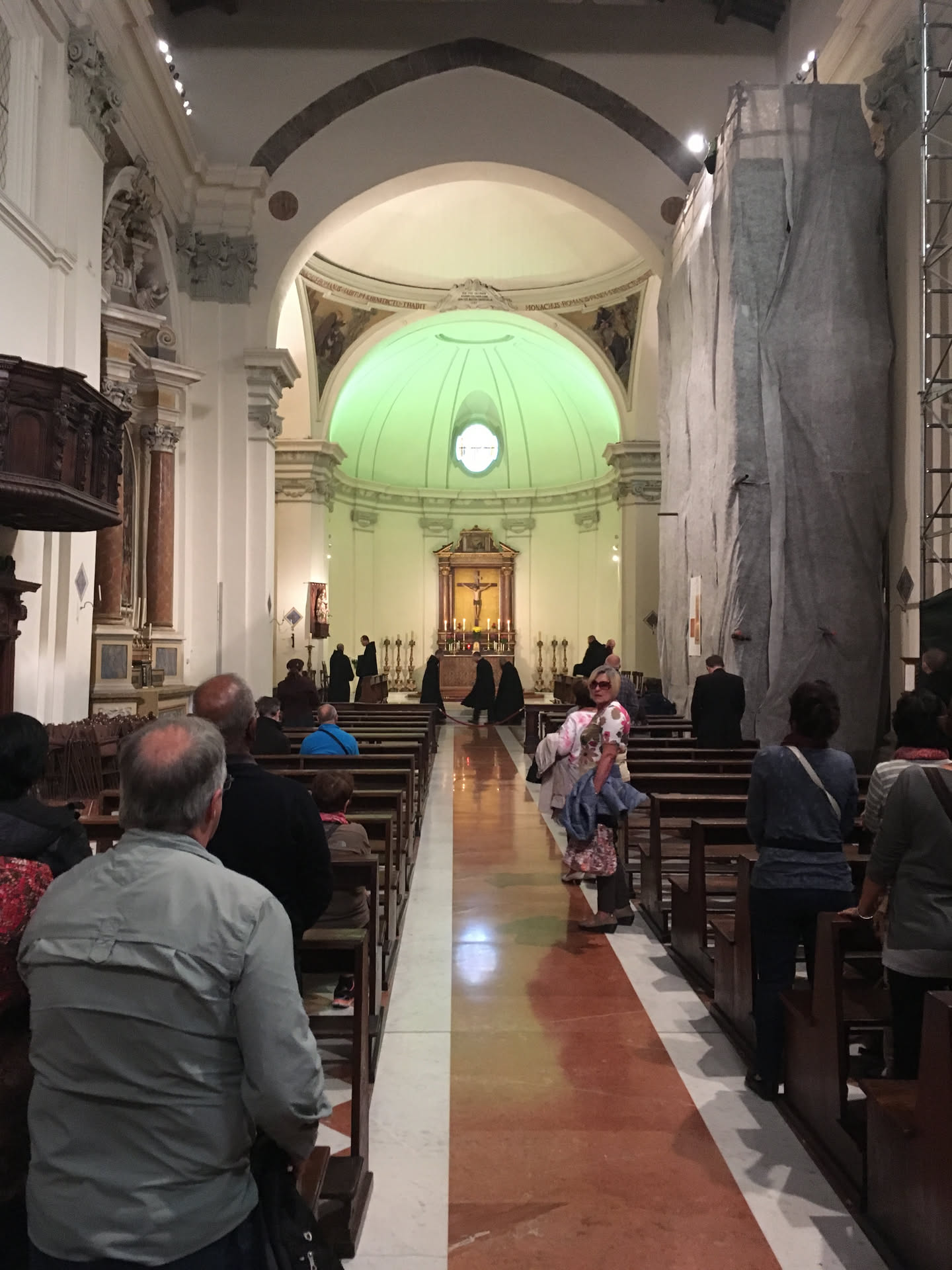 L'interno della basilica nel 2016 durante la messa: i monaci benedettini in cocolla nera davanti all'abside, i fedeli nei banchi, un ponteggio con telo grigio sulla destra
