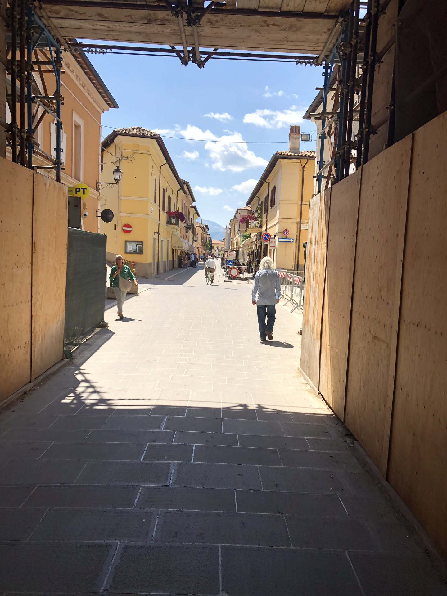 Il corso di Norcia nel 2018 visto da sotto un ponteggio, tra pannelli di compensato; in fondo la strada riaperta con la gente che cammina