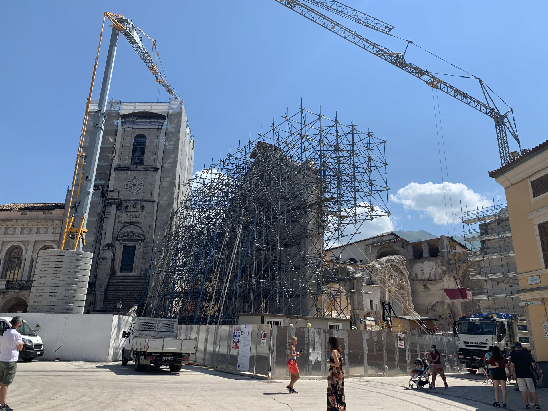 Luglio 2020: due gru a torre sopra la Basilica di San Benedetto, la facciata e il campanile fasciati di ponteggi, la recinzione di cantiere che chiude la piazza