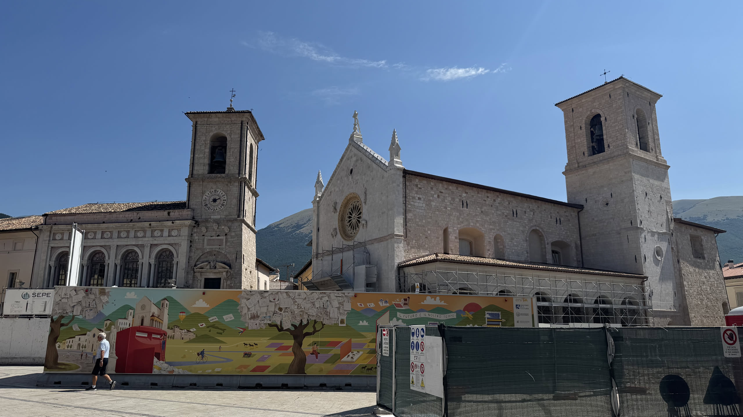 Luglio 2025: la basilica ricostruita, con rosone, tetto e campanile al loro posto, ancora chiusa dietro la recinzione di cantiere dipinta a colori