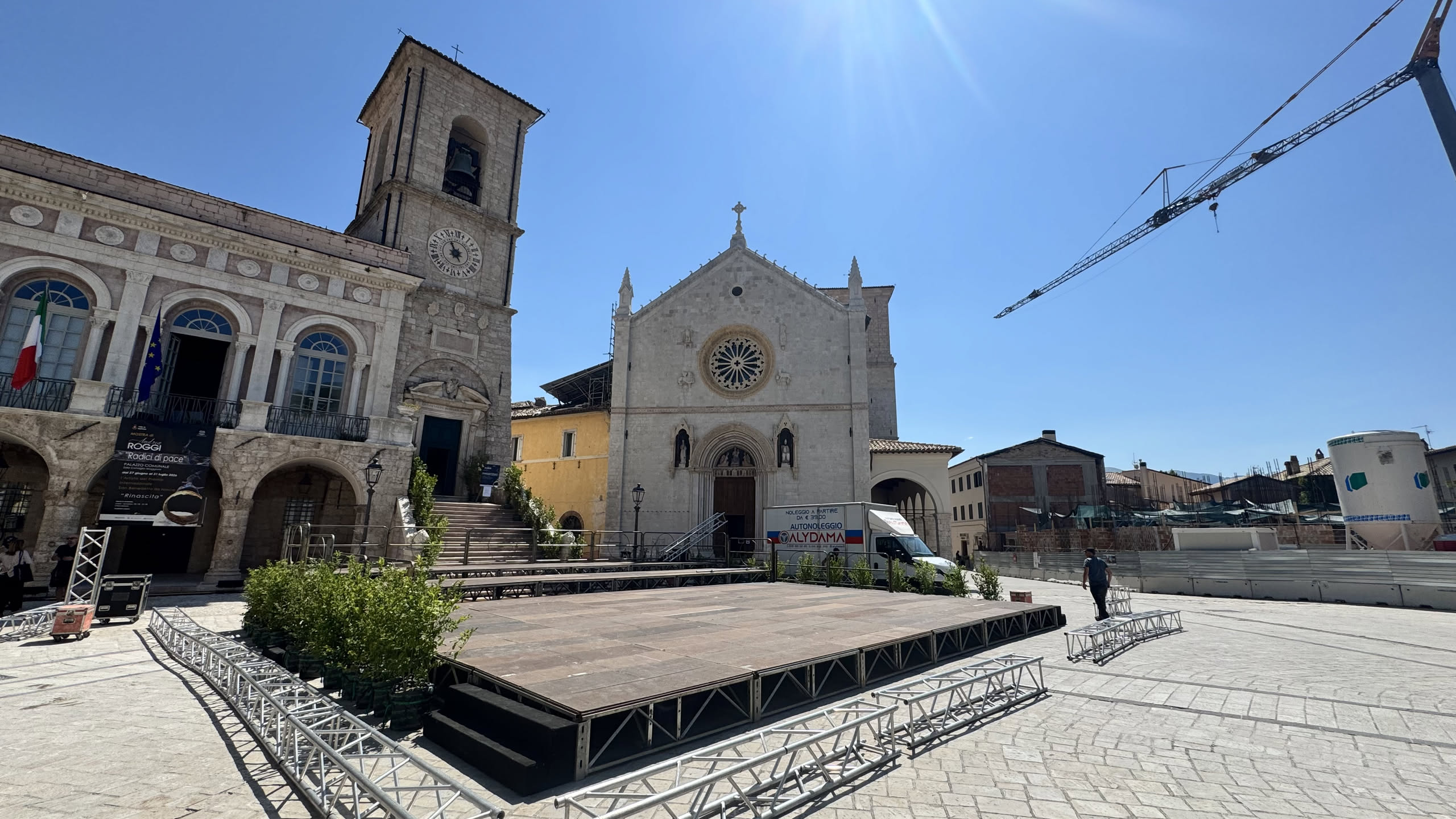 Piazza San Benedetto nel luglio 2026: un palco montato davanti alla basilica aperta, il Palazzo Comunale a sinistra e, sulla destra, la gru di un altro cantiere