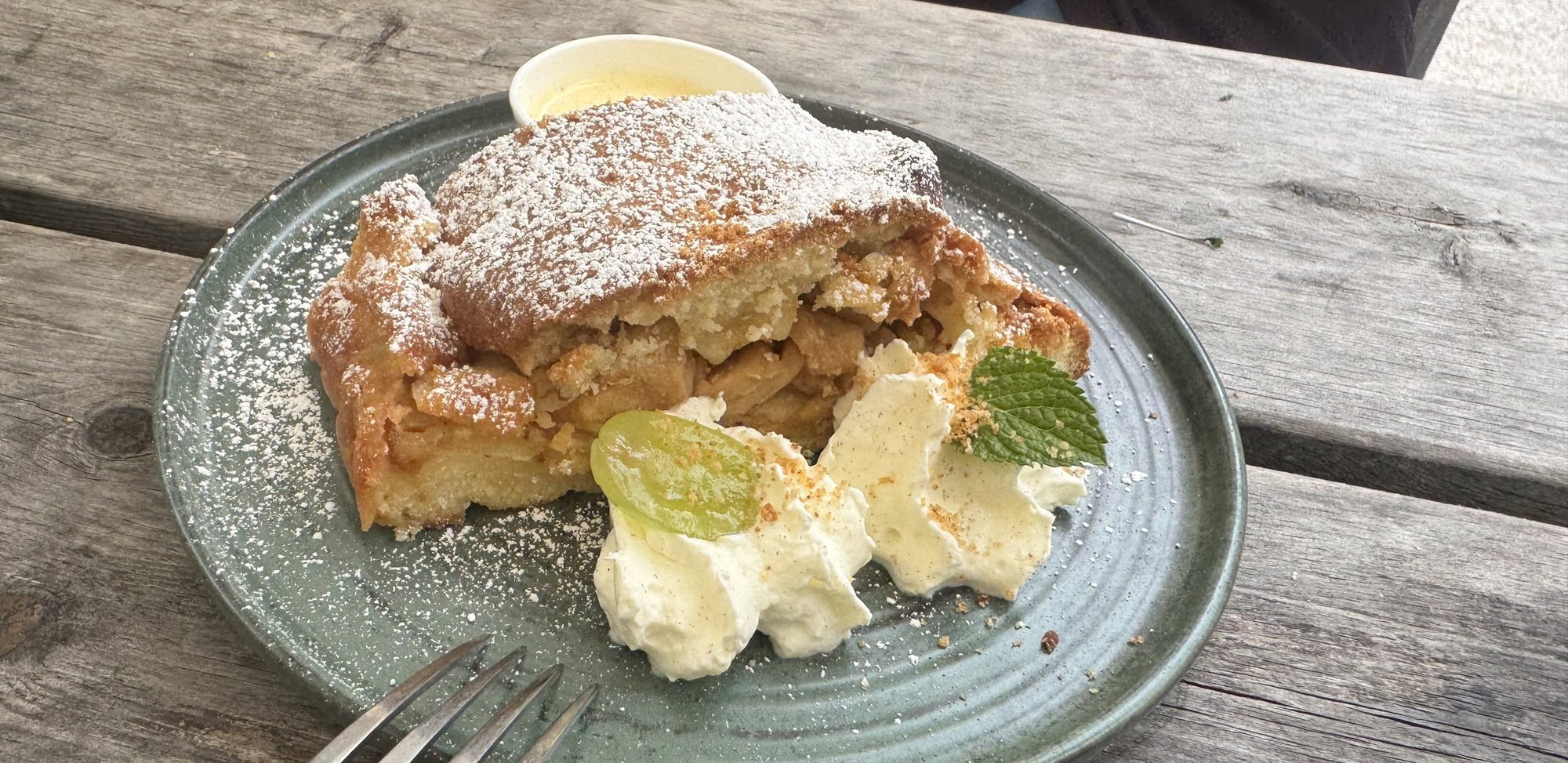 Una fetta di strudel di mele con zucchero a velo, panna e una ciotolina di salsa alla vaniglia, su un piatto verde sul tavolo di legno del rifugio