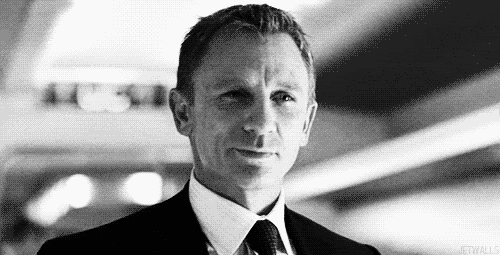 Daniel Craig nei panni di James Bond, in bianco e nero, che sorride e distoglie lo sguardo in un corridoio illuminato