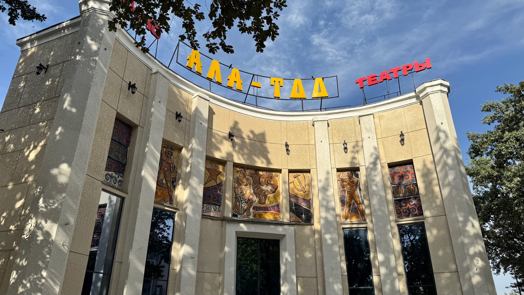 Il cinema Ala-Too: facciata a semicerchio color sabbia, scritta in cirillico rossa e gialla sul tetto e pannelli dipinti fra le colonne