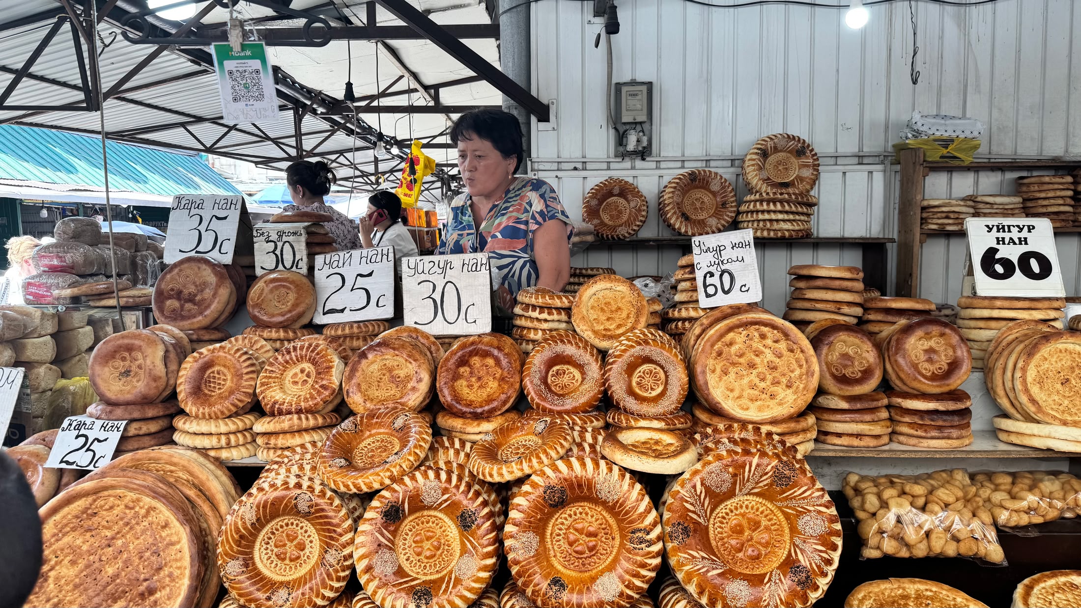 Il banco del pane al bazar: nan tondi stampati col timbro di legno, impilati a torri, con i cartelli dei prezzi da 25 a 60 som