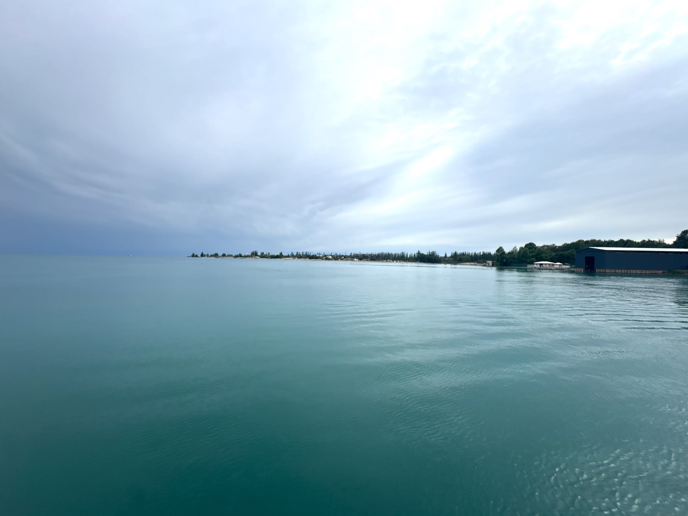 L'acqua turchese dell'Issyk Kul dalla punta del pontile, con la lingua di terra all'orizzonte e il cielo bianco