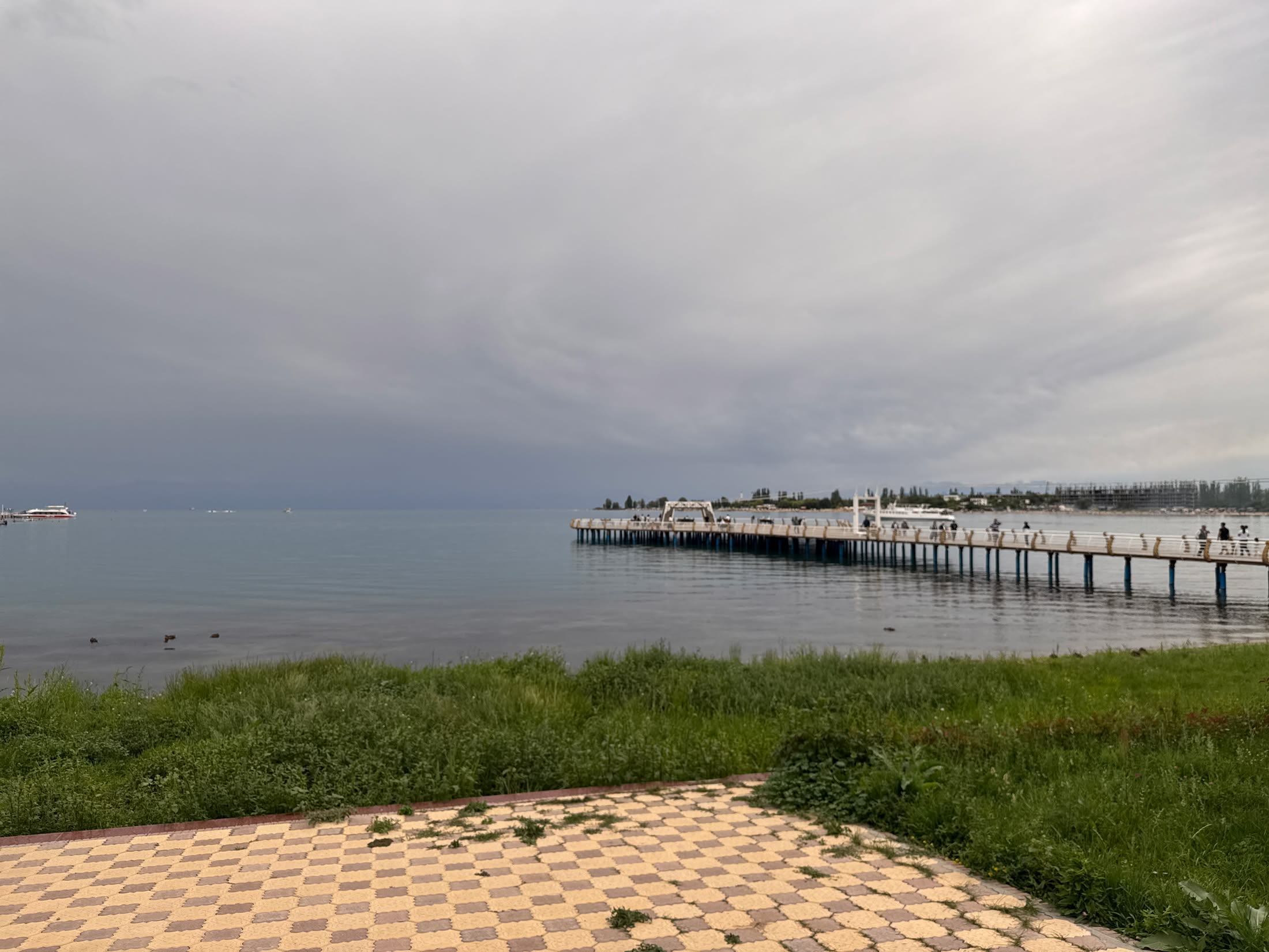 Il lungo pontile di legno che si spinge nell'Issyk Kul sotto un cielo coperto, visto dalla riva pavimentata a scacchi