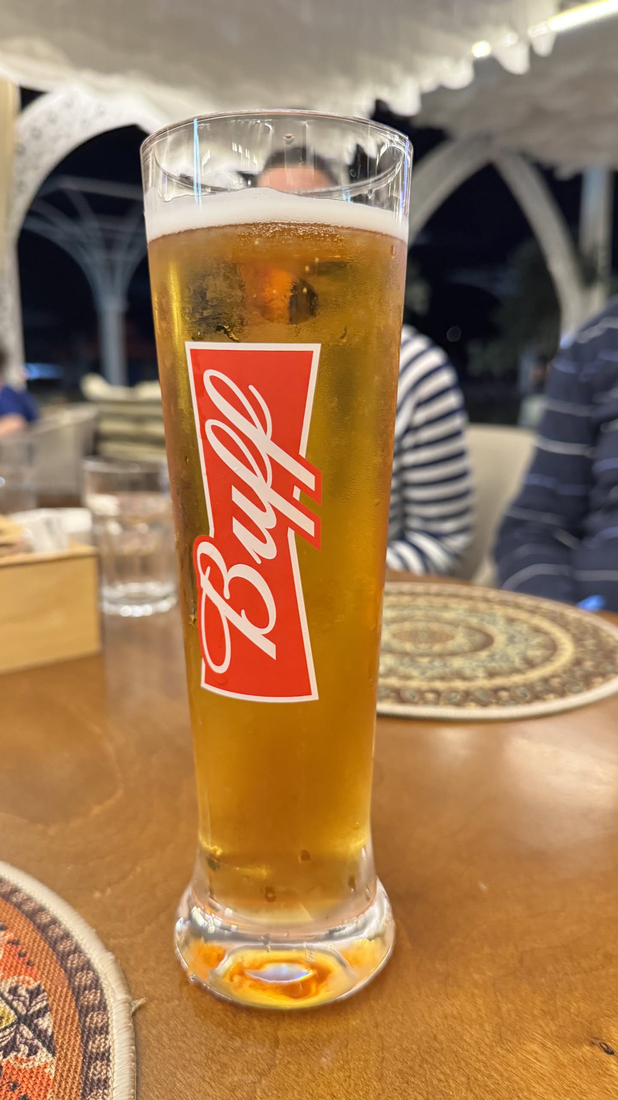 Un bicchiere altissimo di birra chiara sul tavolo di legno, sotto la tenda bianca del ristorante