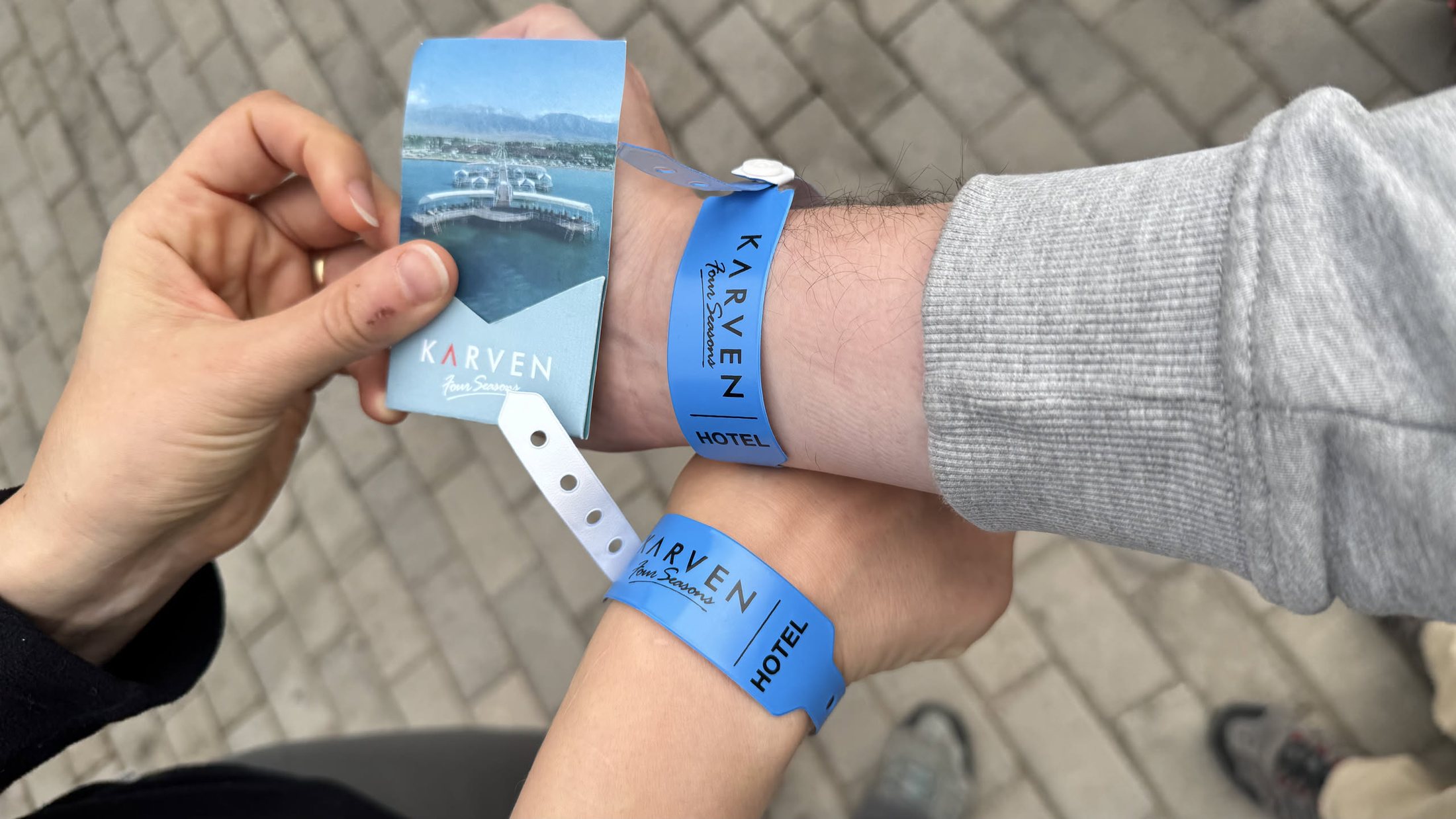 Due polsi con il braccialetto azzurro «KARVEN Four Seasons · HOTEL» e la bustina della chiave con la foto del pontile