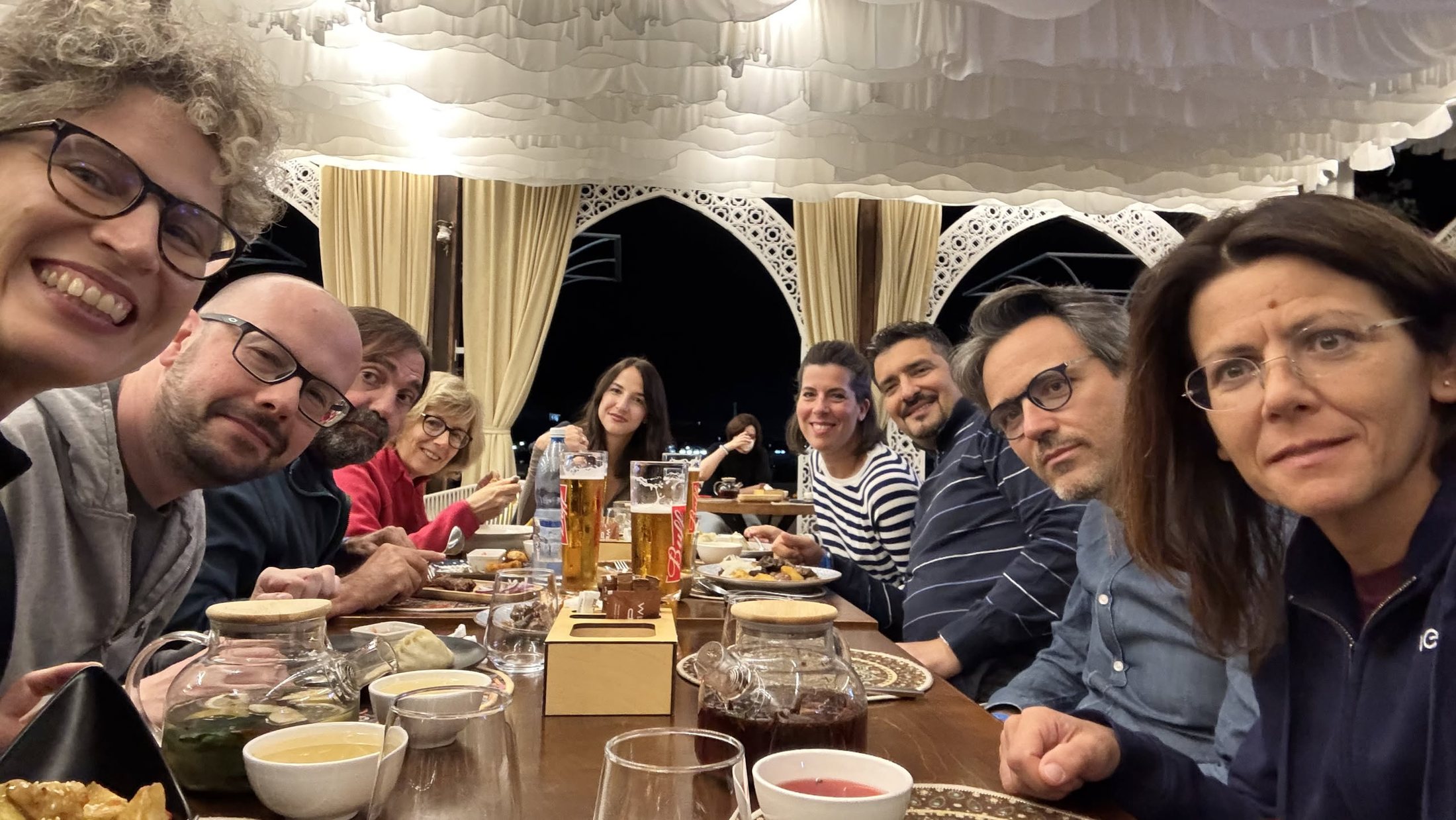 La tavolata del gruppo a cena, dentro il padiglione bianco ad archi, fra ciotole, insalate e bicchieri
