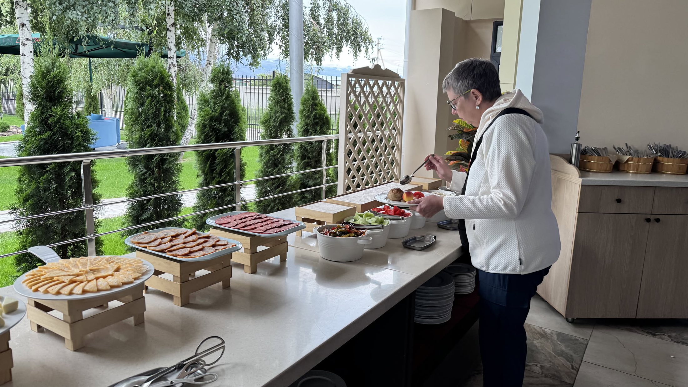 Il buffet della colazione allineato sotto la tettoia, con i cipressi potati sullo sfondo e un signore che si serve