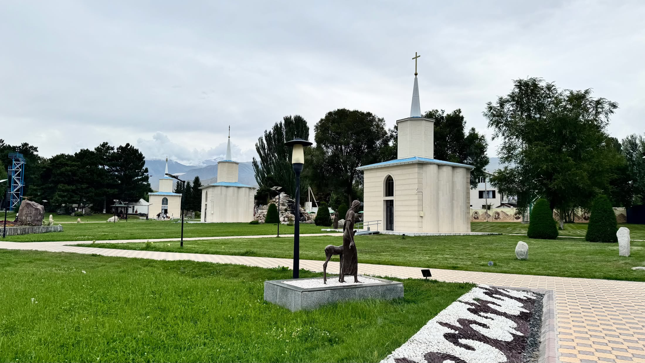 Le cappelle bianche di Rukh Ordo allineate sul prato, con una croce sul campanile e una statua di bronzo lungo il vialetto