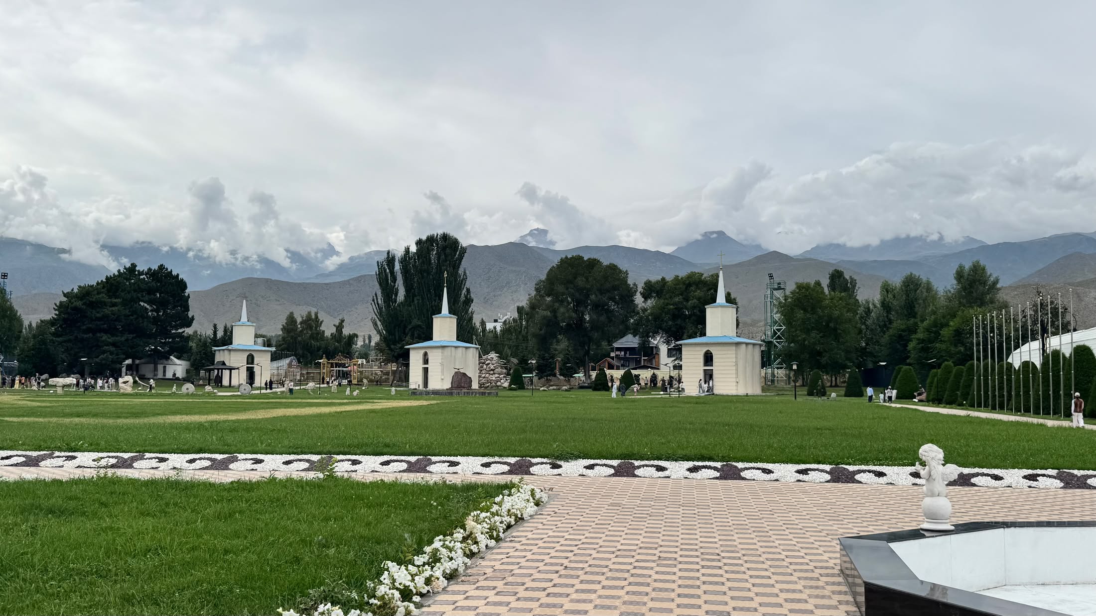 Il complesso di Rukh Ordo dal viale d'ingresso: prato all'inglese, cappelle bianche con le cupole azzurre e le montagne nuvolose dietro